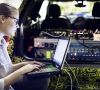 Eine Frau, Mitarbeiterin von BMW, sitzt mit ihrem Laptop vor dem Kofferraum eines Fahrzeugs, in dem elektronische Gerätschaften verkabelt sind.