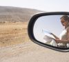 Eine Frau steht mit einer Straßenkarte mitten in der Wüste neben ihrem Auto und ist beim Lesen im Seitenspiegel zu sehen.