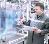Zwei Mitarbeiter von Bosch blicken in einer Produktionshalle auf eine digitla eGrafikillustration, während einer der beiden ein Tablet in der Hand hält.