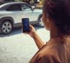 Eine Frau mit lockigem Haar hält ihr SMartphone vor einem Auto in der Hand und nutzt die neue MyMazda App.