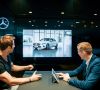 Zwei Männer sitzen an einem Tisch und betrachten ein weißes Fahrzeug von Mercedes-Benz auf einem Bildschirm. Einer der Männer bedient dabei ein Tablet.
